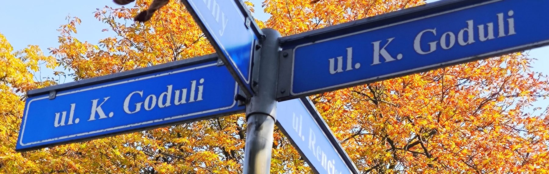 Niebieskie tabliczki z napisem „ul. K. Goduli” na słupie, na tle jesiennych liści
